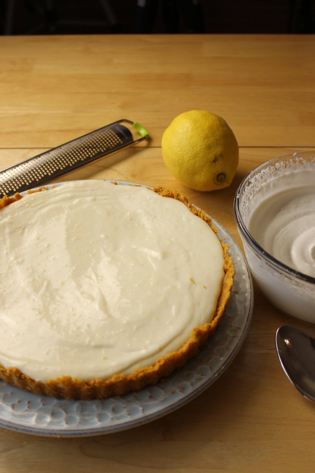 A lemon, a zester, a pie with a creamy filling on a plate, and a bowl of whipped topping sit on a wooden table.