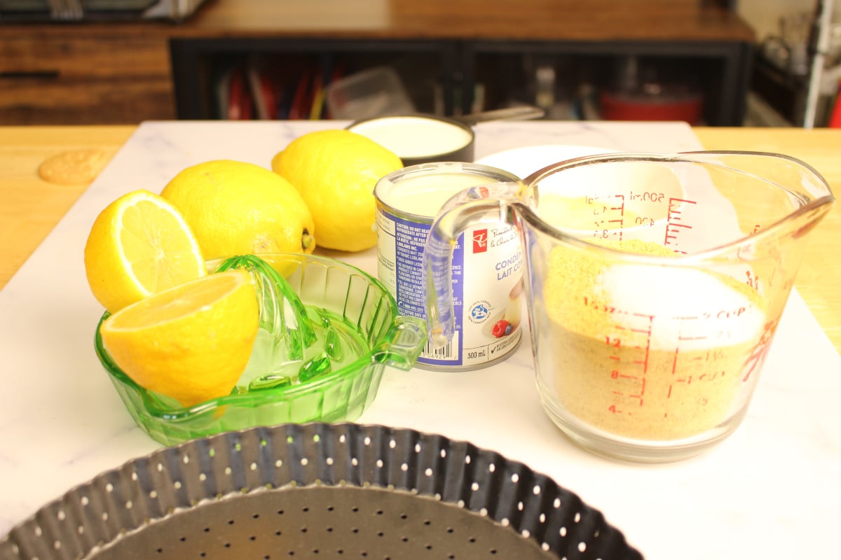 Lemons, a glass lemon juicer, a can of sweetened condensed milk, a measuring cup of sugar, and an empty tart pan on a countertop.