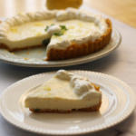 A slice of cream pie with a graham cracker crust and whipped topping sits on a plate; the rest of the pie is on a serving dish in the background.