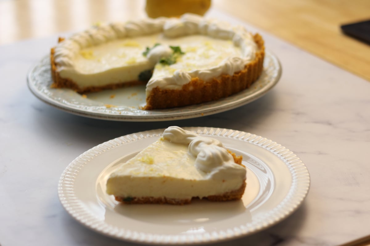 A slice of cream pie with a graham cracker crust and whipped topping sits on a plate; the rest of the pie is on a serving dish in the background.
