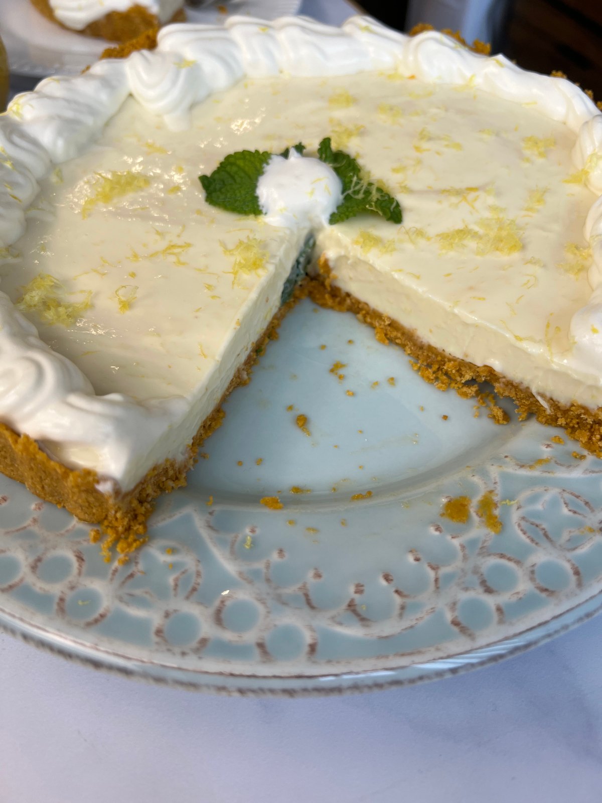 A creamy lemon pie with a graham cracker crust, topped with whipped cream, lemon zest, and a mint leaf, served on a decorative plate with one slice missing.