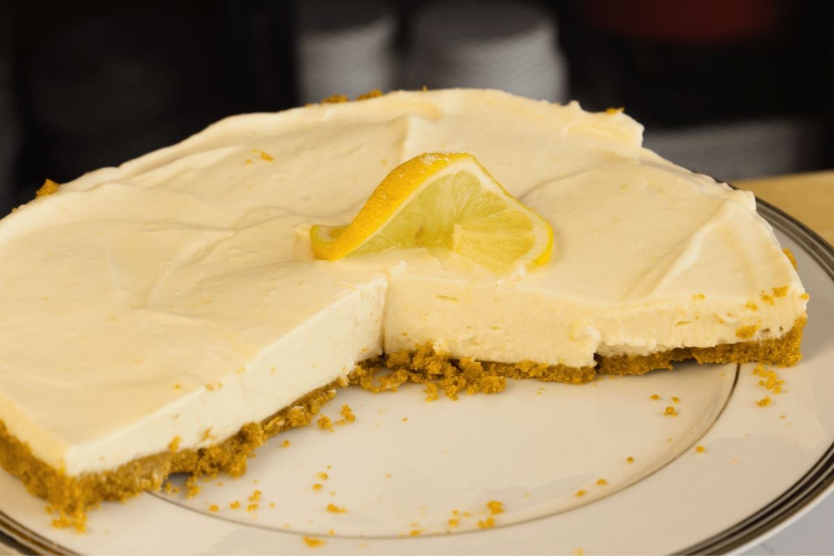 A cheesecake with a graham cracker crust and a slice missing, topped with a lemon wedge, resembles an easy no bake lemon tart, beautifully displayed on a plate.
