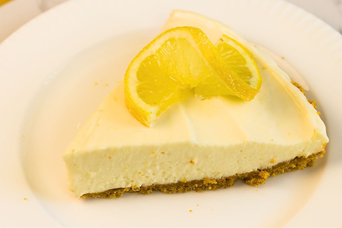 A slice of lemon cheesecake with a graham cracker crust, topped with two lemon slices, served on a white plate—an easy lemon dessert no bake fans will love.