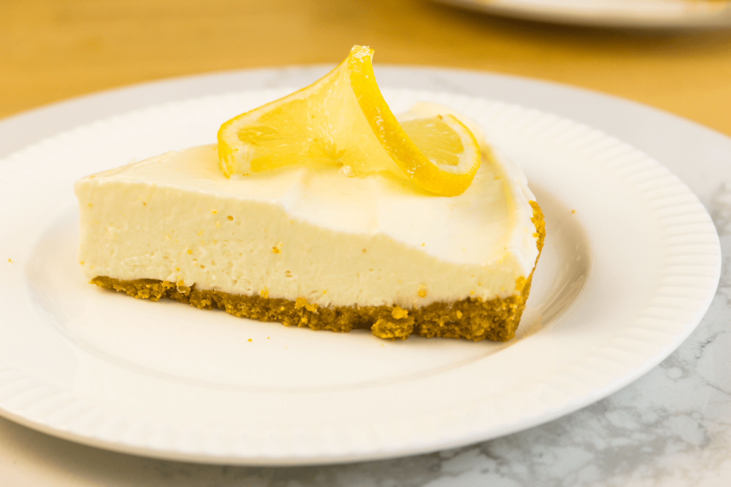 A slice of lemon cheesecake with a graham cracker crust, inspired by an easy lemon tart recipe, garnished with two lemon slices and served on a white plate.