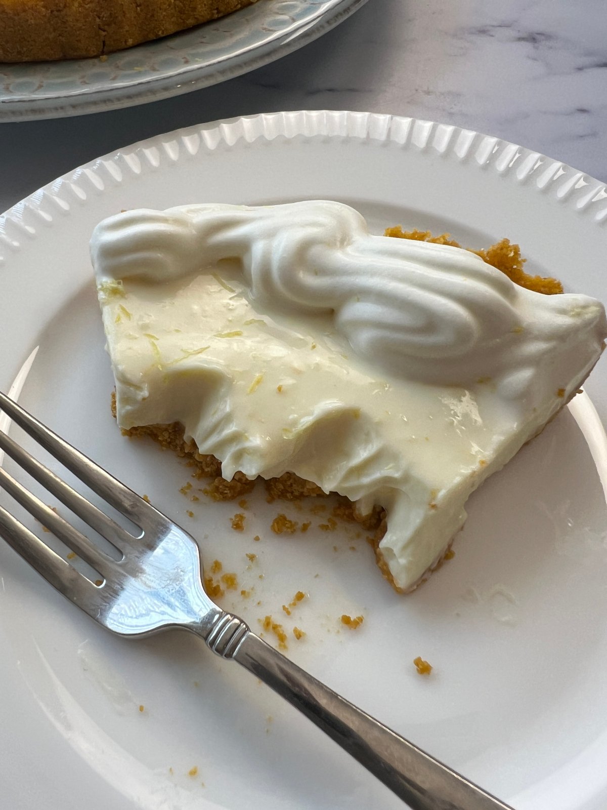 A partially eaten slice of creamy pie with a graham cracker crust and whipped topping on a white plate with a fork.