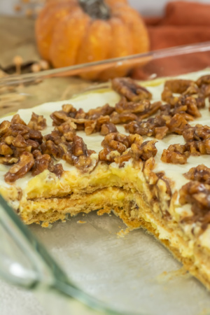 A glass dish containing a layered dessert topped with glazed walnuts, with one large piece missing; a pumpkin is visible in the background.