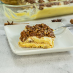 A square serving of layered dessert with a pecan topping sits on a white plate, with a glass baking dish and scattered spices in the background.