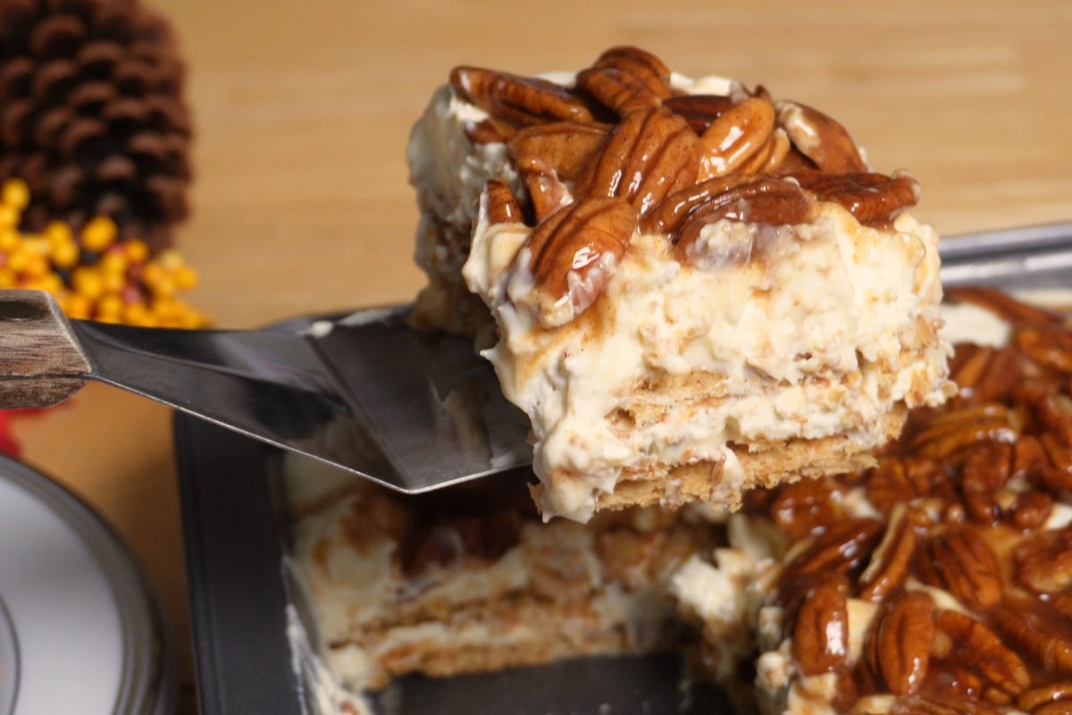 A metal spatula lifts a square slice of layered pecan pie lasagna cheesecake from a baking pan.