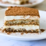 A slice of tiramisu with layers of coffee-soaked cake and creamy filling, topped with a dusting of cocoa powder, served on a white plate.