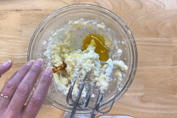 A hand holds a glass bowl with softened butter, sugar, vanilla, and egg being mixed by electric beaters on a wooden surface.