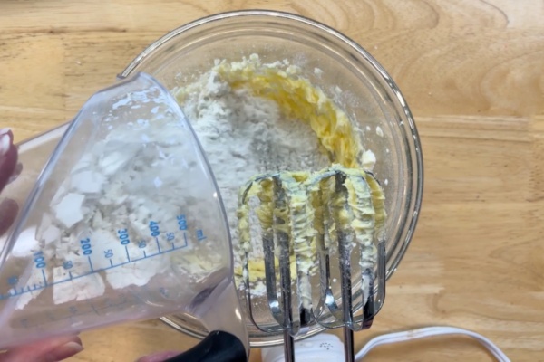 A person pours flour from a measuring cup into a glass bowl containing butter and sugar, with an electric hand mixer in use on a wooden surface.
