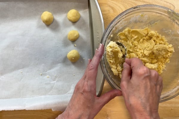 Hands shaping cookie dough and placing it on a parchment-lined baking sheet, with a bowl of dough and spoon on the right side.