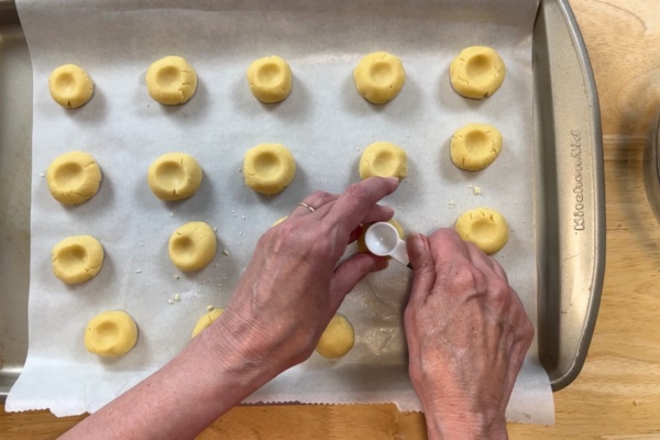 Hands pressing indentations into rows of unbaked cookie dough balls on a parchment-lined baking sheet.