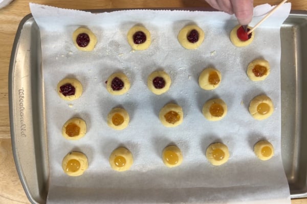 A baking tray lined with parchment paper holds rows of thumbprint cookies being filled with red and orange jams; a hand uses a toothpick to add jam to one cookie.