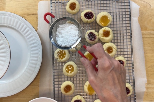 A hand sprinkles powdered sugar over jam-filled thumbprint cookies on a cooling rack, with plates stacked nearby.