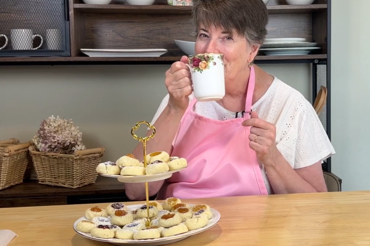 An older woman in a pink apron drinks from a floral mug, sitting at a table with a two-tier tray of cookies in a cozy kitchen setting.
