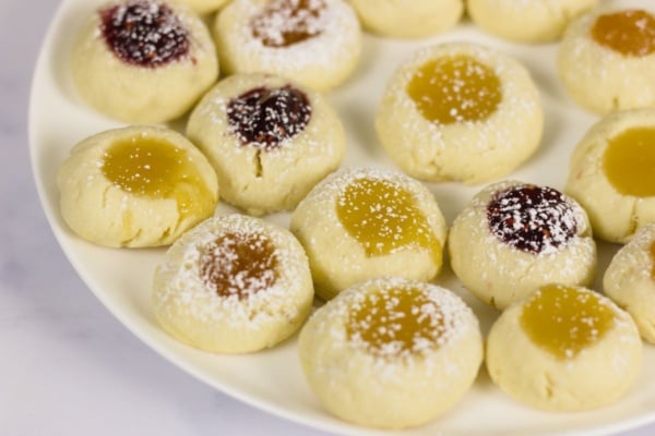 A plate of thumbprint cookies filled with yellow and red jam, some dusted with powdered sugar, arranged on a white plate.