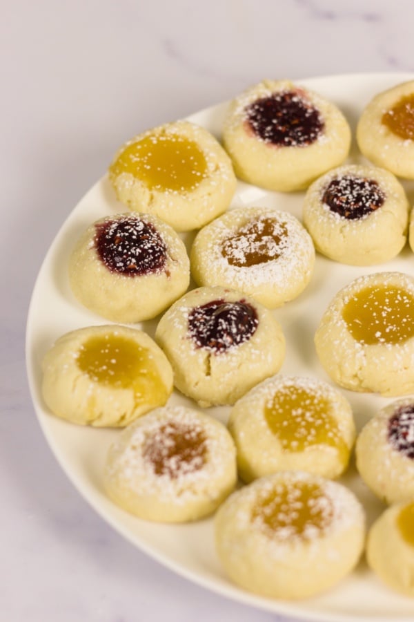 A plate of thumbprint cookies filled with yellow and dark red jam, dusted with powdered sugar, on a white surface.
