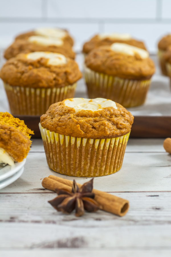Banana muffins with banana slices on top are arranged on a wooden surface, with cinnamon sticks and star anise in the foreground.