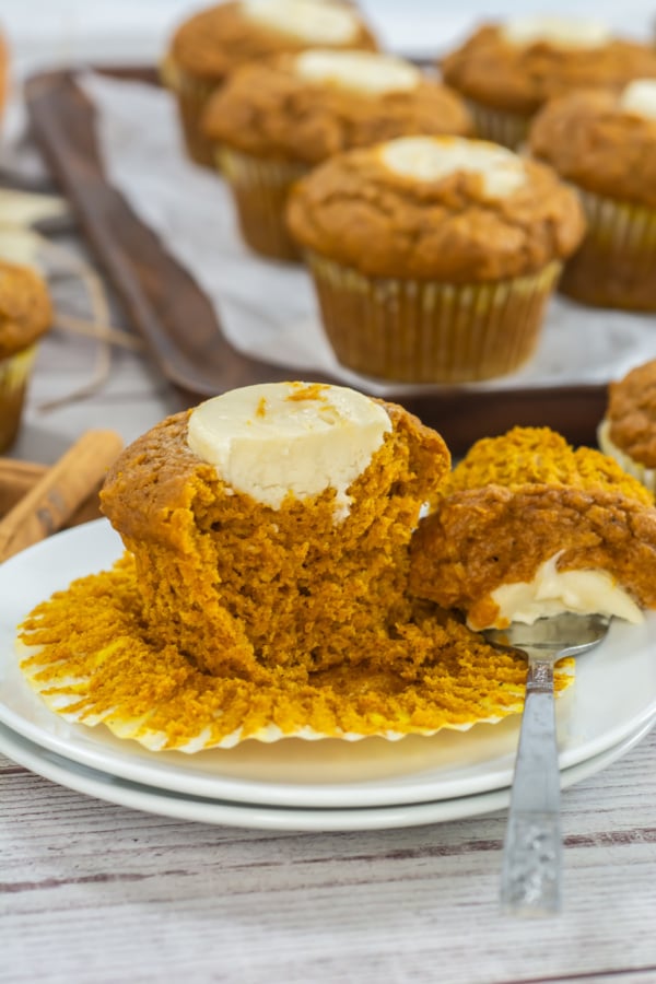 A pumpkin muffin with cream cheese filling sits on a plate, partially unwrapped and cut with a spoon. More muffins are on a tray in the background.