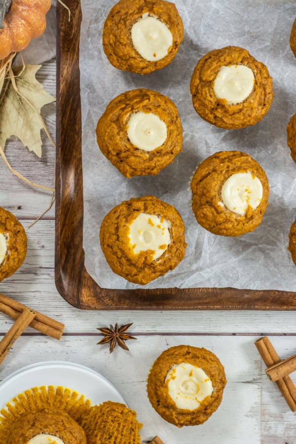 Pumpkin muffins with cream cheese centers arranged on a wooden tray lined with parchment paper, with spices and leaves nearby.
