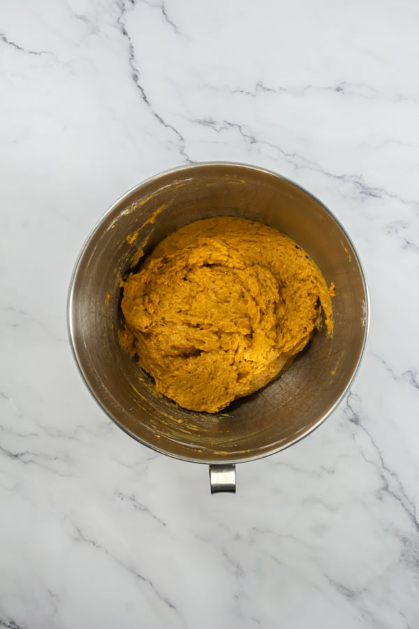 A stainless steel mixing bowl containing orange-colored dough sits on a white marble surface.