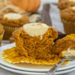 A Starbucks Pumpkin Cream Cheese Muffin is split open on a plate, revealing its creamy filling, surrounded by similar muffins on a table.