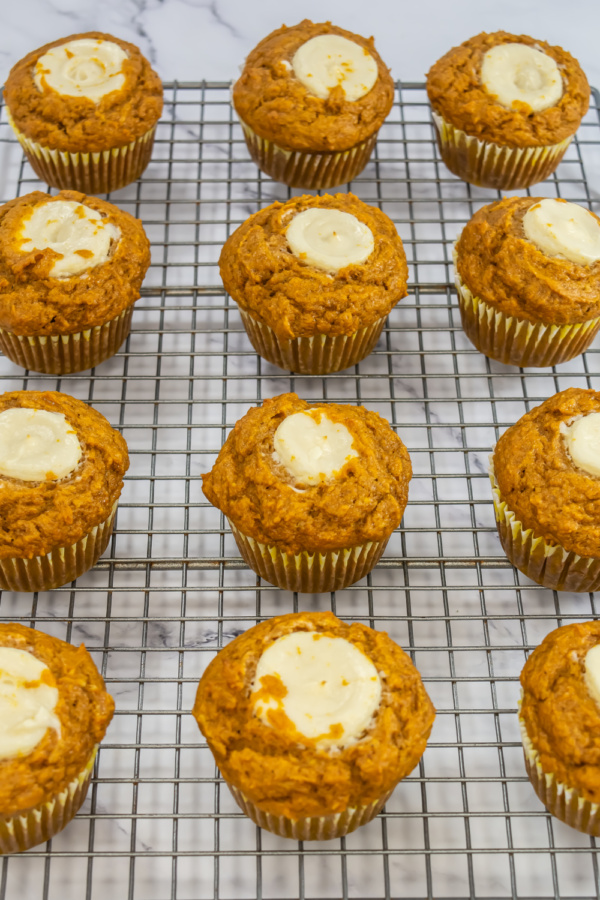 Twelve pumpkin muffins with cream cheese centers are arranged on a cooling rack over a marble surface.