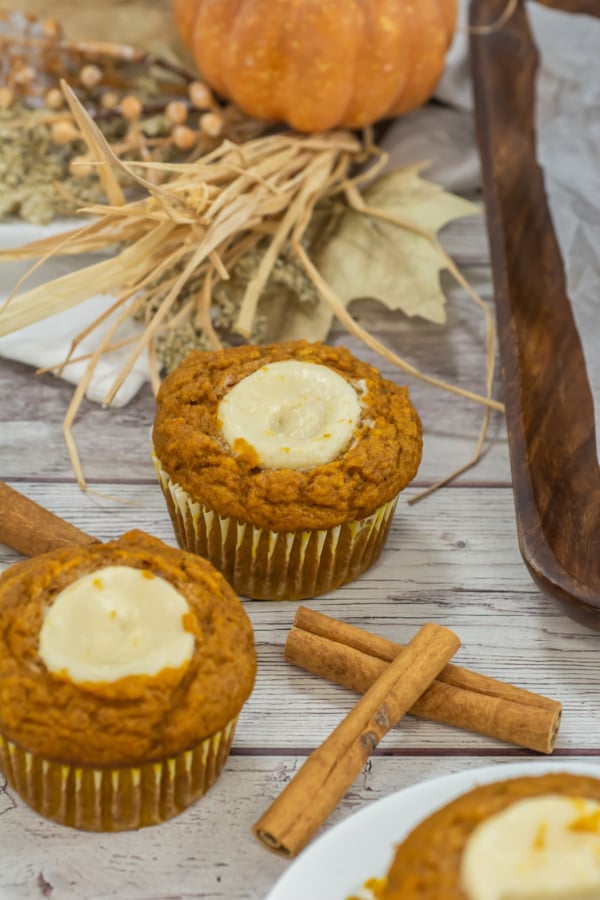 Two pumpkin muffins with cream cheese centers sit on a wooden table, surrounded by cinnamon sticks, autumn leaves, and a small pumpkin in the background.