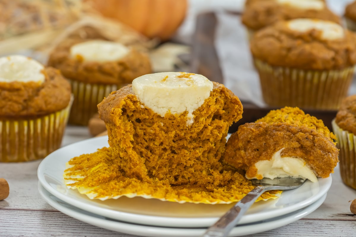 A Starbucks Pumpkin Cream Cheese Muffin is split open on a plate, revealing its creamy filling, surrounded by similar muffins on a table.