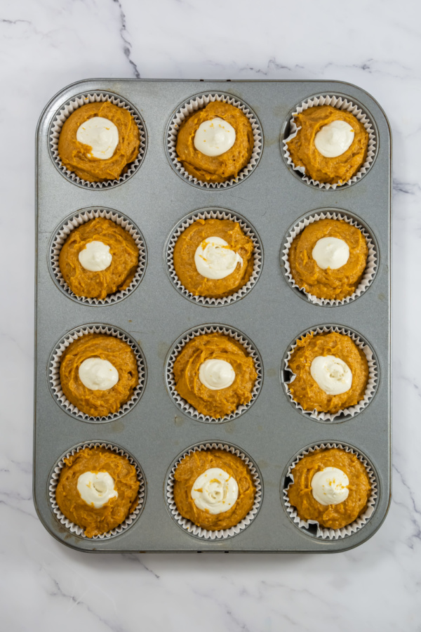 A muffin tin with twelve unbaked Starbucks Pumpkin Cream Cheese Muffins, each topped with a small dollop of cream cheese, sits on a marble countertop.
