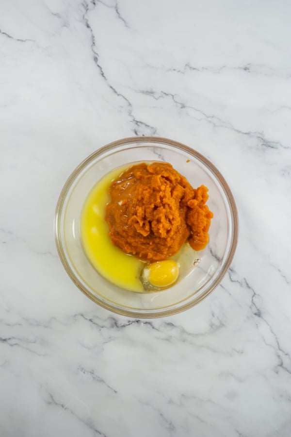 A glass bowl containing pumpkin puree, a cracked raw egg, and melted butter on a white marble surface—just the start for homemade Starbucks Pumpkin Cream Cheese Muffins.