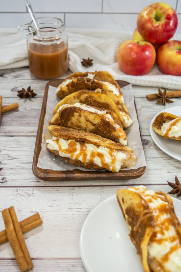 Apple cinnamon dessert tacos filled with cream and drizzled with caramel sauce are arranged on a wooden tray, with apples, caramel sauce, and cinnamon sticks in the background.