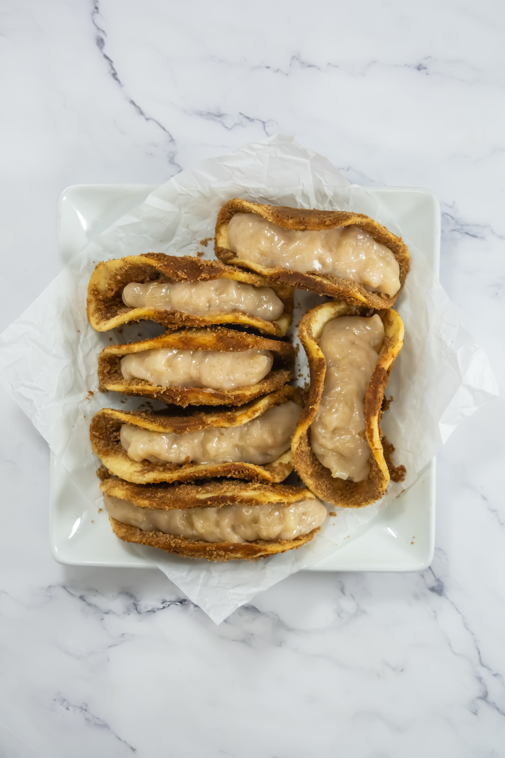 A square white plate with parchment paper holds seven pieces of pie crust filled with a creamy, light beige filling on a marble surface.