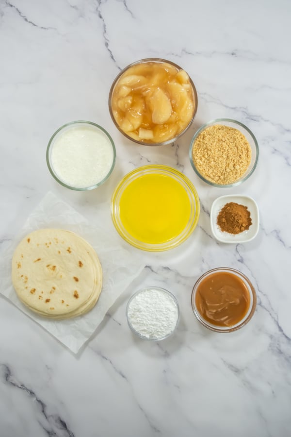 Flat lay of tortillas, canned apple pie filling, melted butter, crushed graham crackers, ground cinnamon, caramel sauce, powdered sugar, and milk on a marble surface.