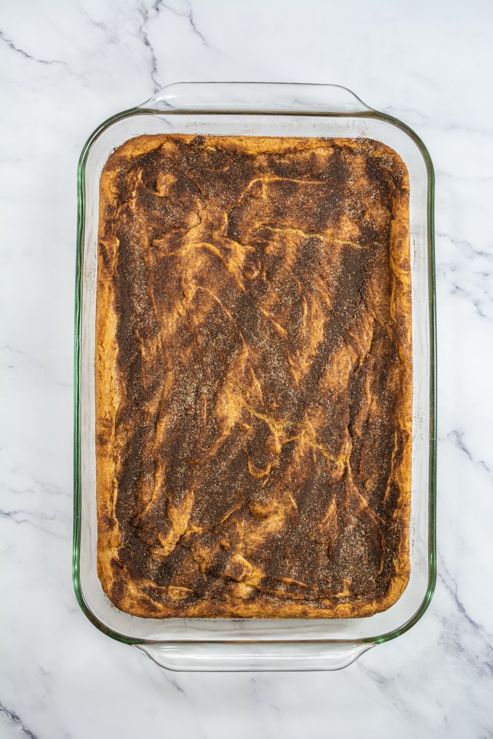 A glass baking dish filled with a baked dessert featuring a golden-brown, cinnamon-sugar topped surface on a white marble background.