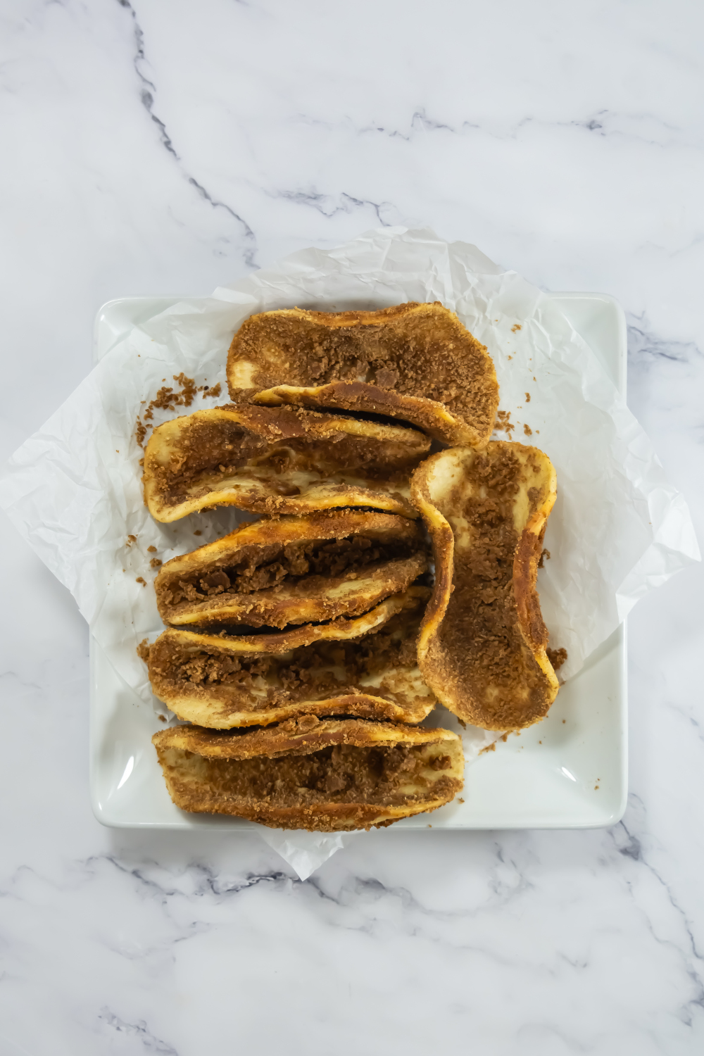 A square white plate with six pieces of cinnamon toast on white parchment paper, viewed from above on a marble surface.