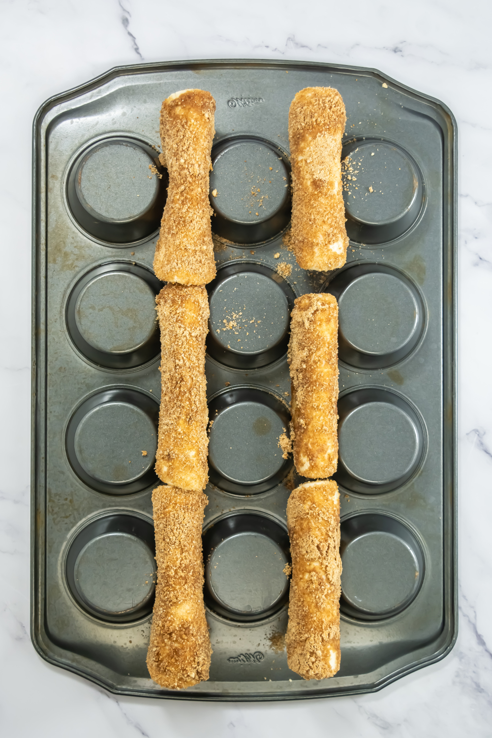 Six coated breadsticks arranged across the tops of an inverted muffin pan, ready for baking, on a marble surface.