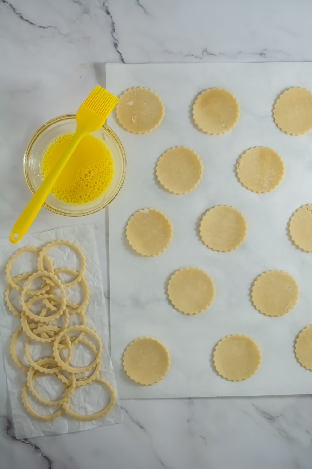 Round pastry dough circles are arranged on a marble surface next to cut dough rings, a bowl of beaten egg, and a yellow pastry brush.