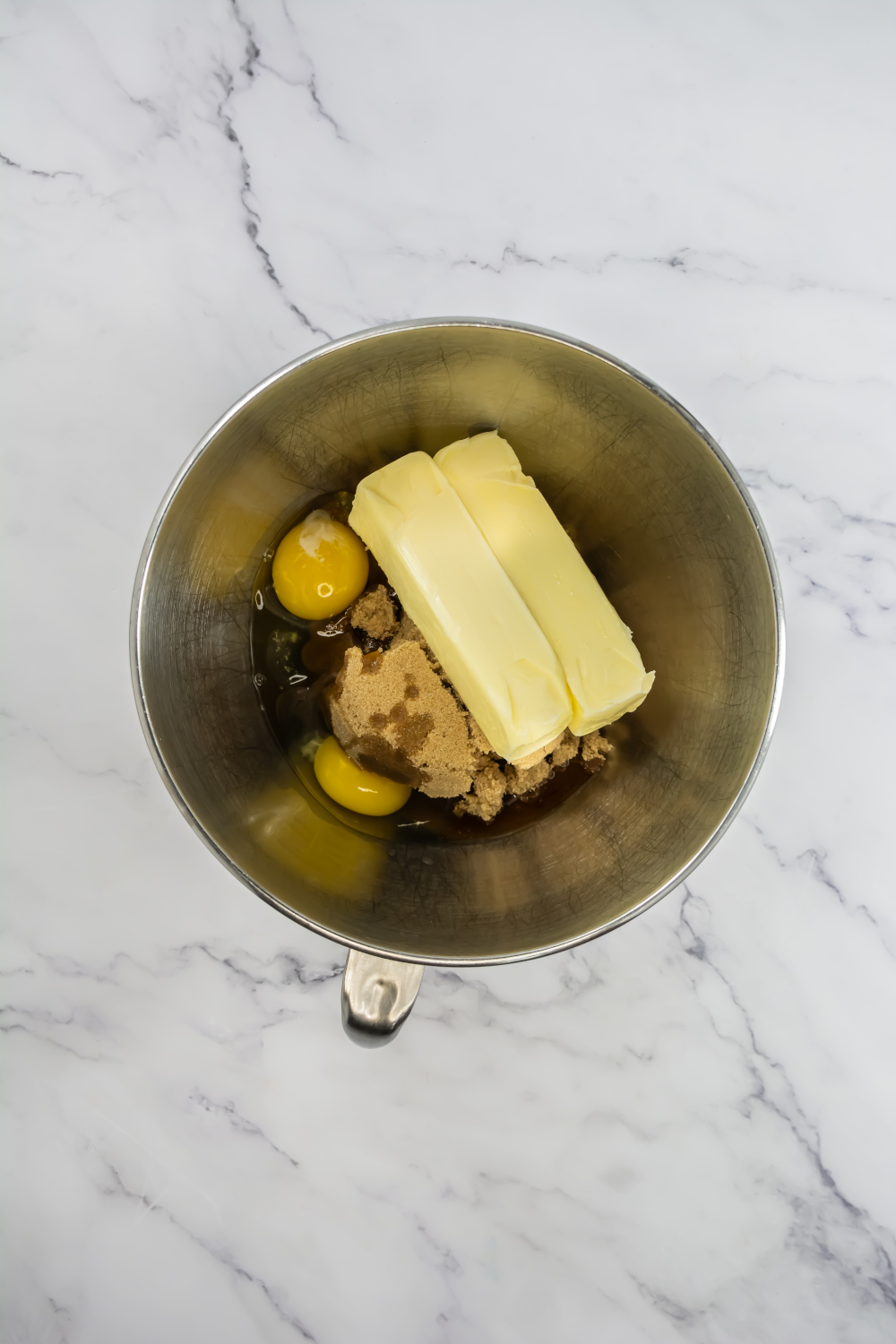 A metal mixing bowl contains two eggs, two sticks of butter, brown sugar, and vanilla extract on a white marble surface.