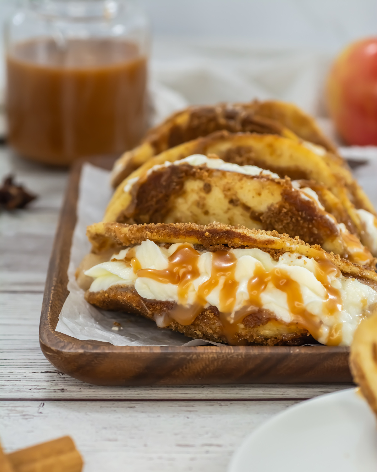 A row of dessert tacos filled with cream and topped with caramel sauce on a wooden tray, with a jar of caramel and an apple in the background.