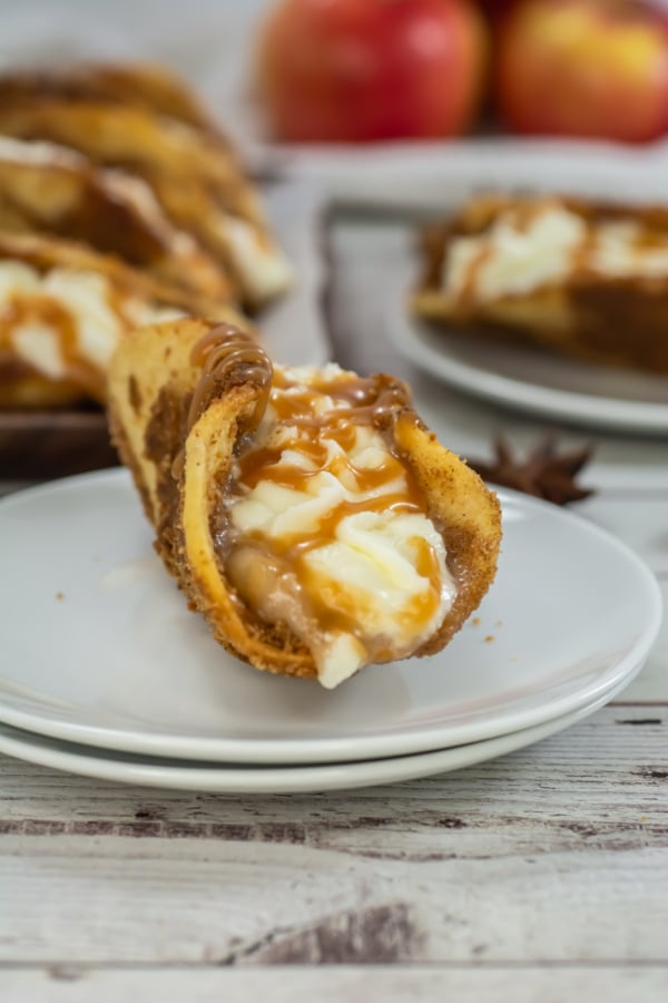 A dessert taco with a cookie shell filled with cream and apple filling, topped with caramel sauce, is placed on a white plate with apples in the background.