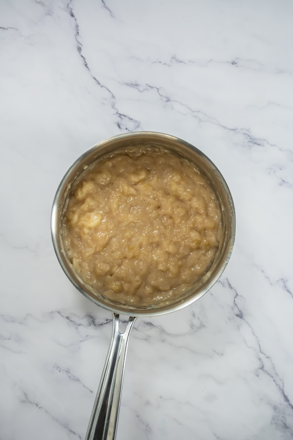A metal saucepan containing mashed bananas sits on a white marble countertop.