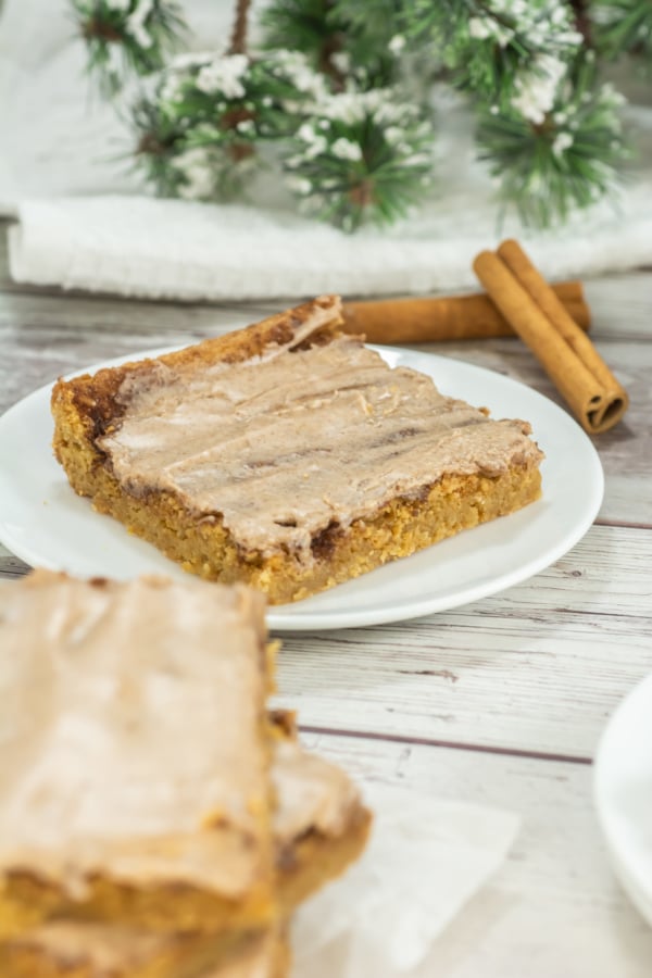 A frosted bar slice on a white plate sits on a light wooden table with cinnamon sticks and snowy evergreen decor in the background.