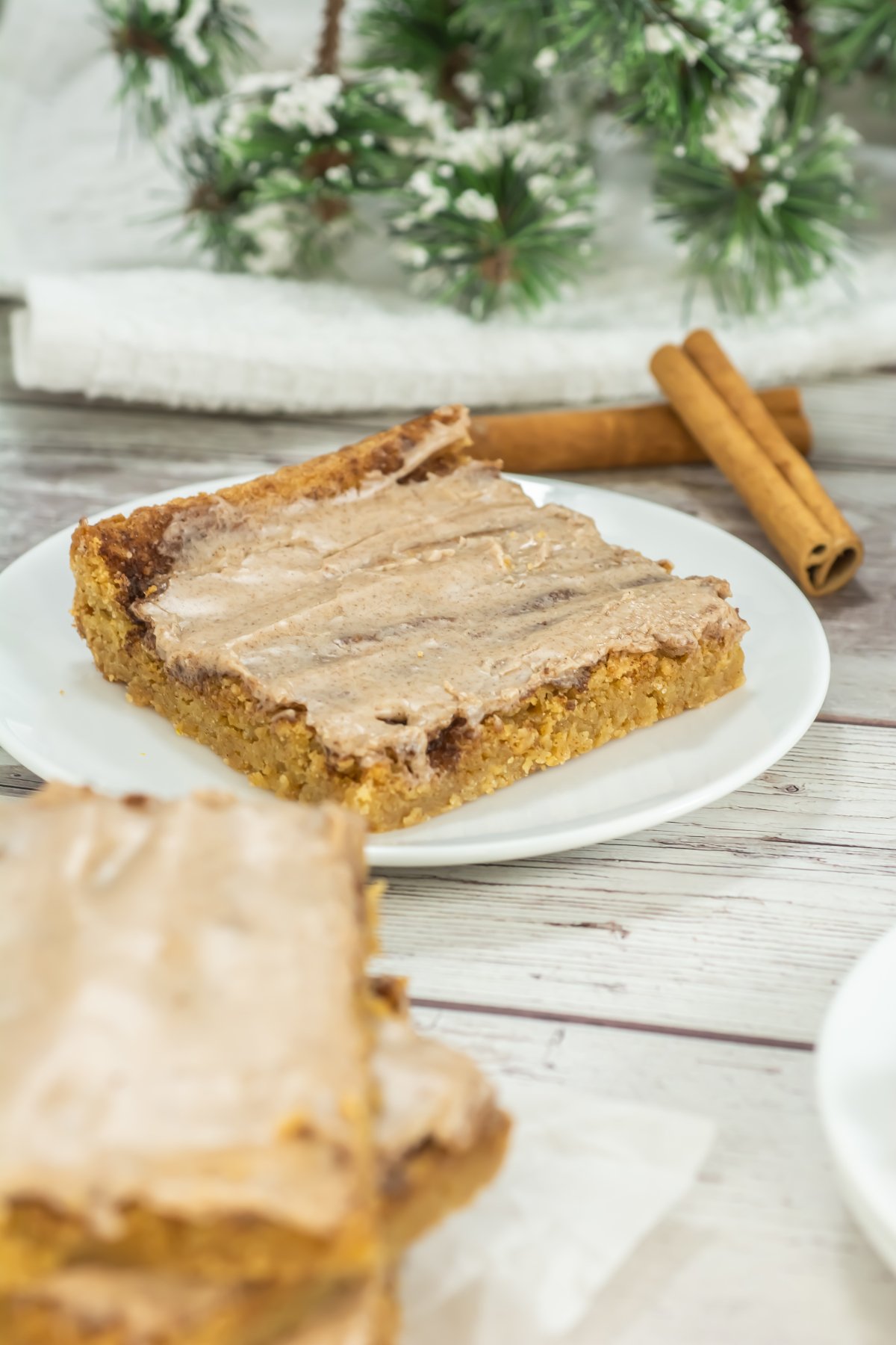 A frosted bar slice on a white plate sits on a light wooden table with cinnamon sticks and snowy evergreen decor in the background.