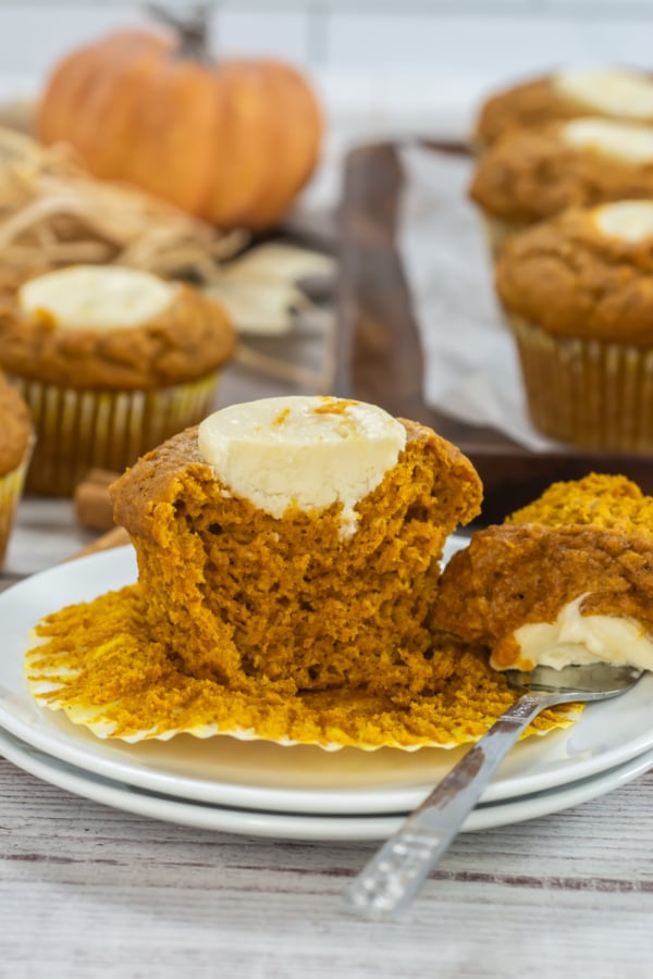 A pumpkin muffin with cream cheese filling is cut open on a plate with a fork; more muffins and a pumpkin are in the background.