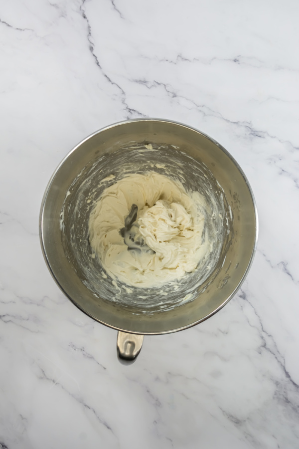 A metal mixing bowl containing whipped cream cheese mixture sits on a white marble surface.