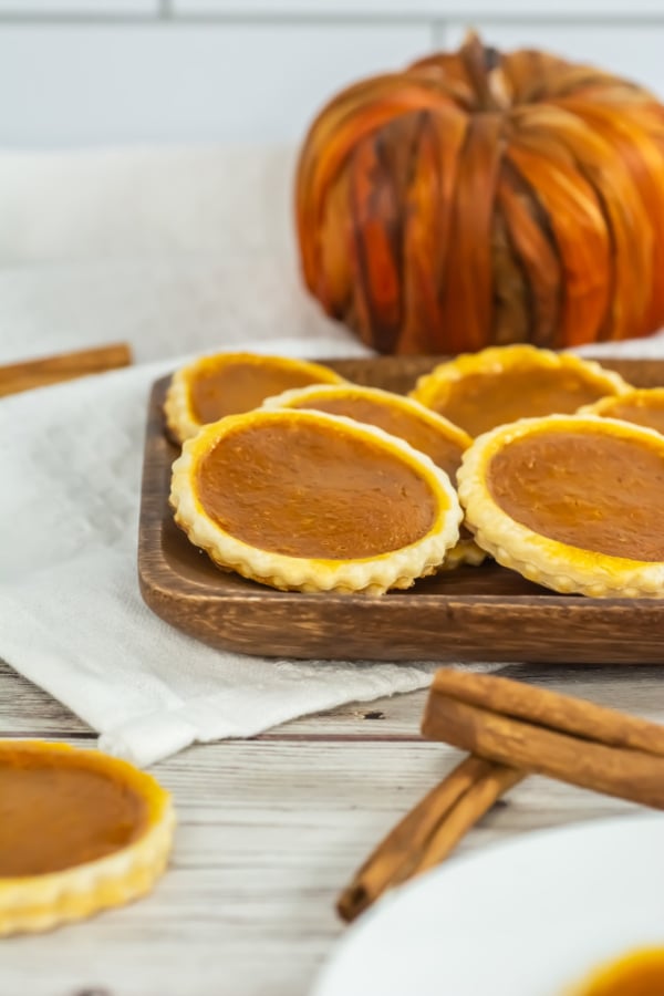 Mini pumpkin pies arranged on a wooden tray with cinnamon sticks nearby and a decorative pumpkin in the background.