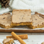 Three frosted cinnamon bars are arranged on a wooden tray lined with parchment paper, with cinnamon sticks placed in front and a white towel in the background.
