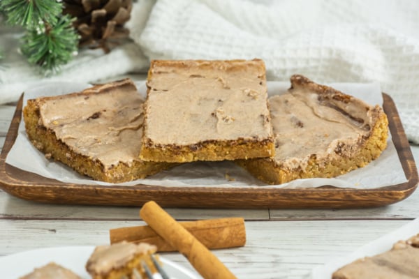 Three frosted cinnamon bars are arranged on a wooden tray lined with parchment paper, with cinnamon sticks placed in front and a white towel in the background.